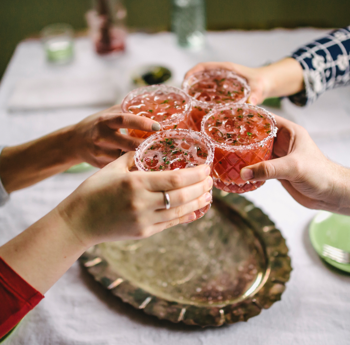 Four hands clink glasses filled with a pink beverage, garnished with herbs, over a silver tray on a white tablecloth, celebrating new beginnings in Peoria AZ.