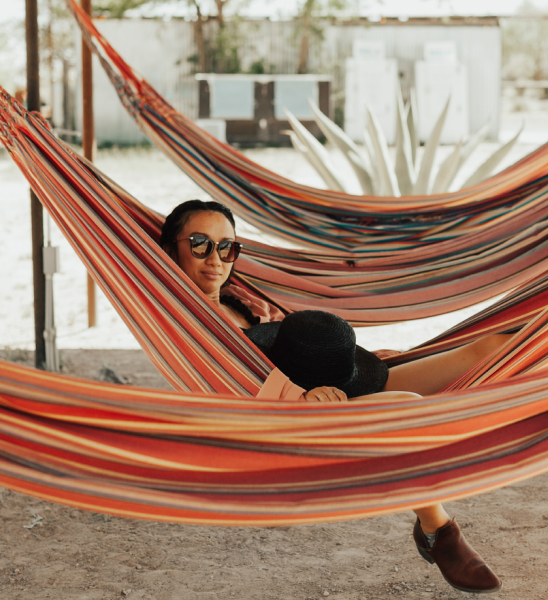 A person relaxes in a striped hammock outdoors under the Saddleback sun in Peoria, AZ, wearing sunglasses and holding a dark hat.