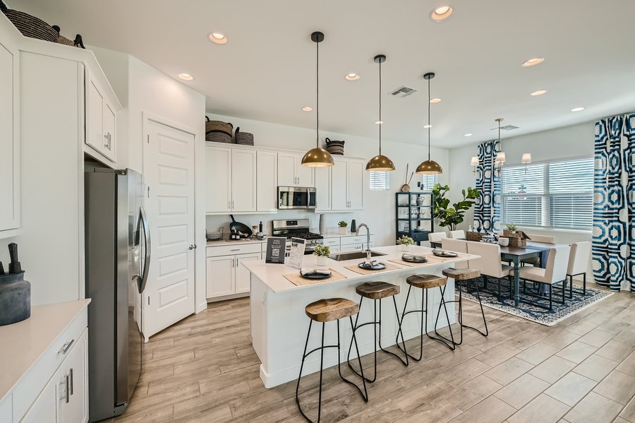Modern kitchen with white cabinets, stainless steel appliances, and a central island with wooden bar stools. Dining area with a table and patterned curtains in the background. Hanging pendant lights.