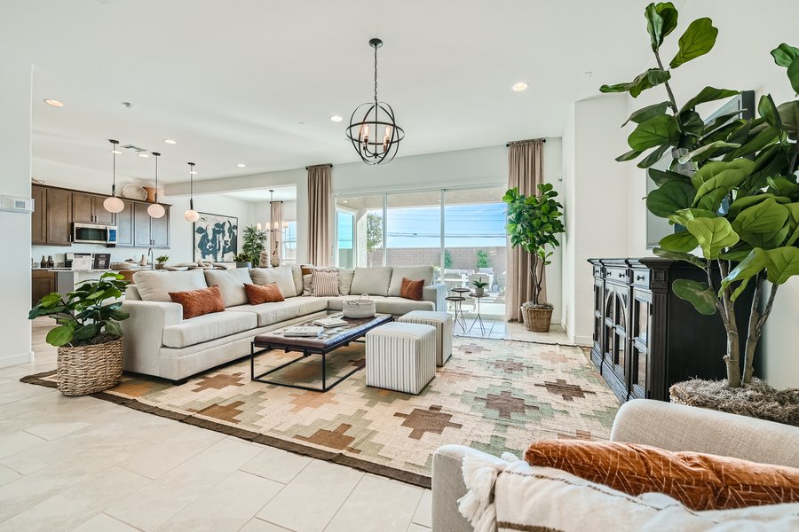 Spacious living room with a large sectional sofa, geometric rug, and plants. A modern chandelier hangs from the ceiling. Open view into the kitchen and glass doors lead to an outdoor area.