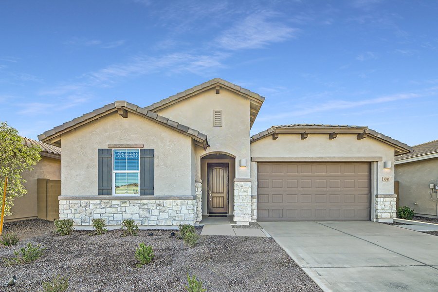 Single-story beige house with stone accents, two-car garage, and a small front yard. The sky is clear and blue.