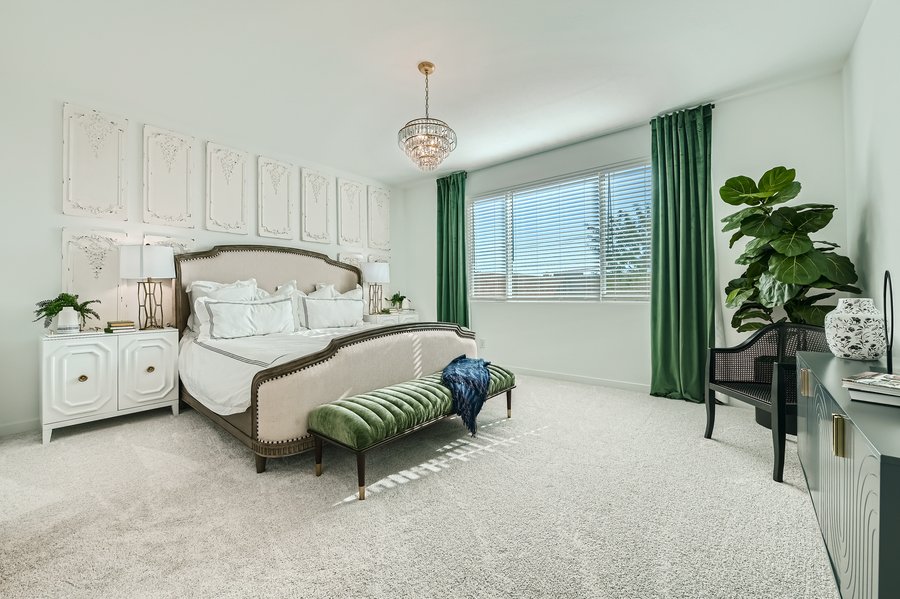Spacious bedroom with a large bed, green curtains, and a crystal chandelier. A green bench sits at the foot of the bed. A window with blinds lets in natural light.