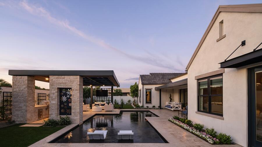 A modern backyard at dusk with a rectangular pool, patio area, outdoor kitchen, and seating. The house exterior is light-colored with large windows and landscaped with flowers.