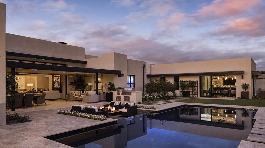 Modern house exterior at dusk, featuring a lit outdoor seating area, pool, and large windows displaying indoor living spaces. Clouds and a soft evening sky are visible in the background.