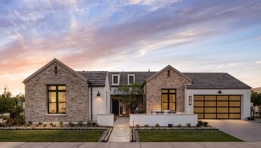 Modern single-story house with two brick facades, large windows, and a double garage. Manicured lawn with a pathway leads to the entrance. Sky features a vibrant sunset.