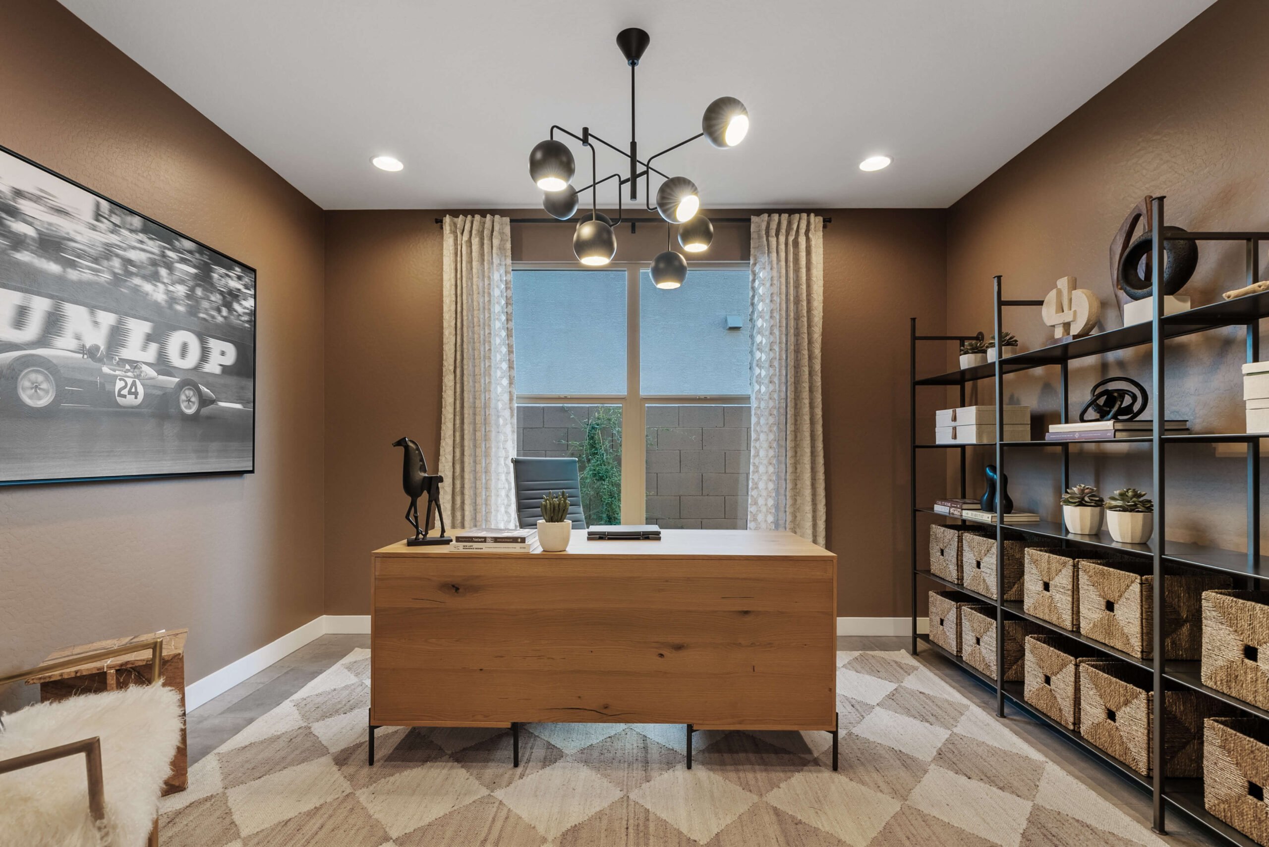 A modern home office with a wooden desk, geometric rug, and shelving unit with woven baskets. A large black-and-white art piece and contemporary light fixture add flair to the brown-toned space.