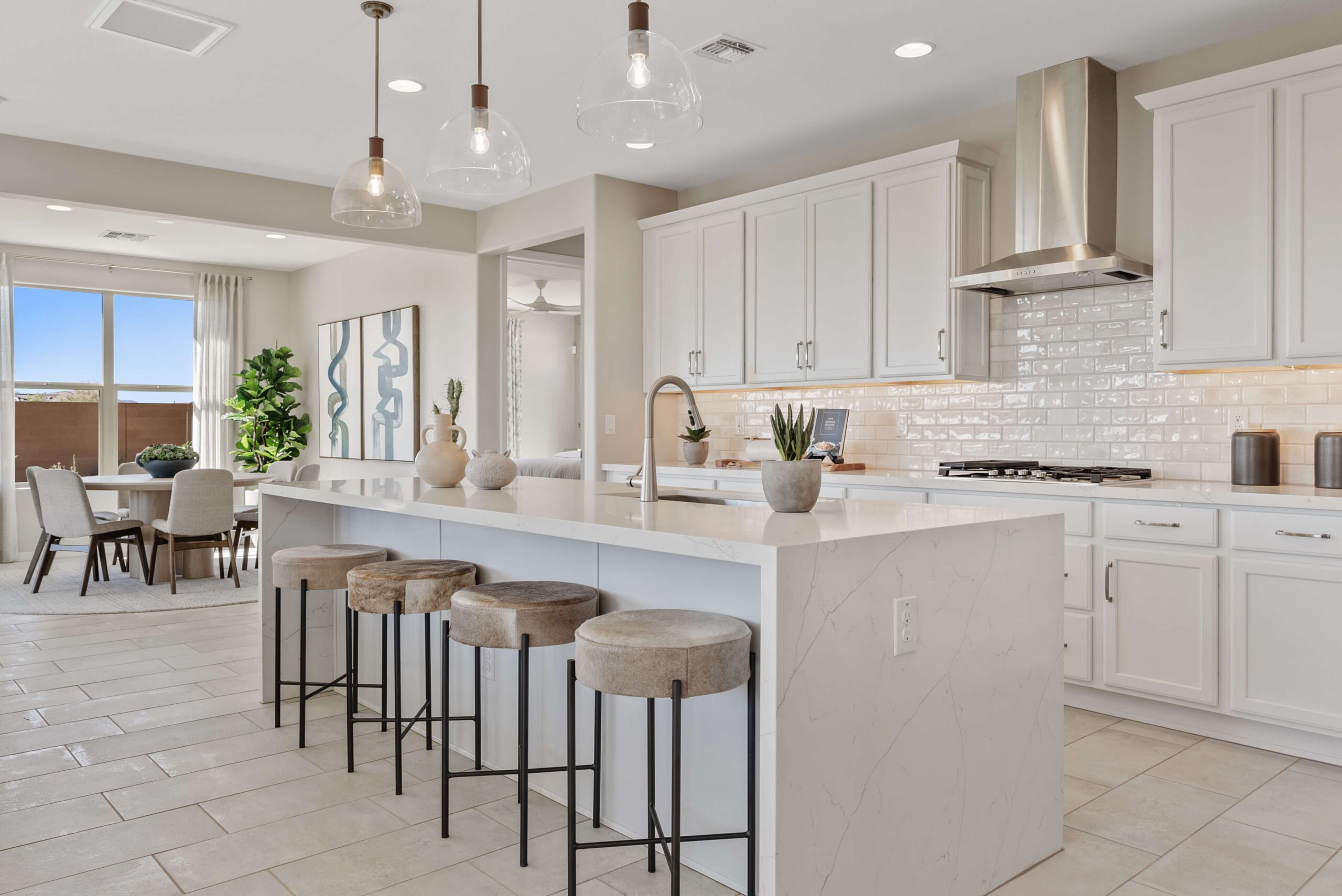 Modern kitchen with a large central island, bar stools, white cabinetry, stainless steel appliances, and pendant lighting. Dining area with table and chairs is visible in the background.