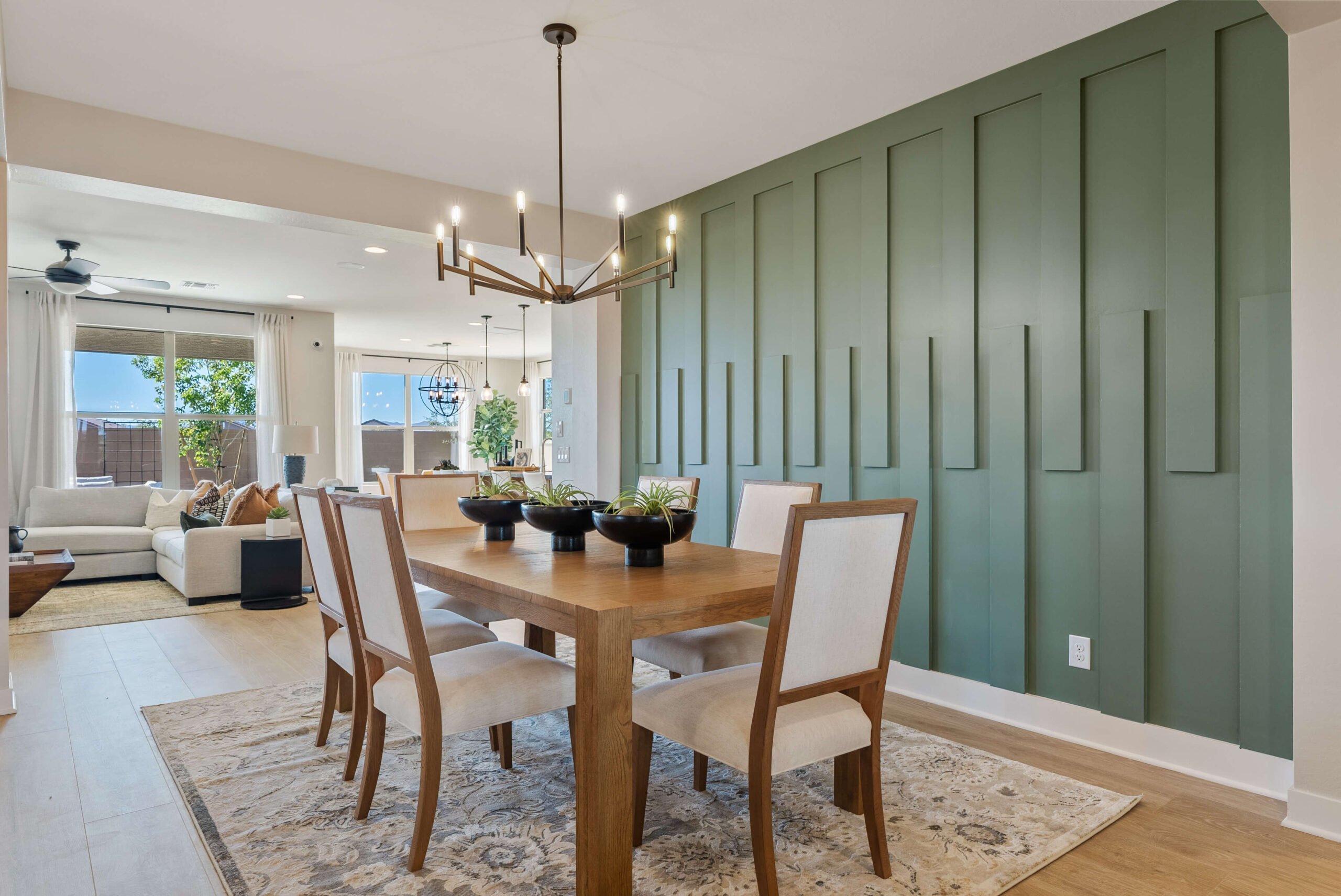 Dining area with a wooden table and six chairs, set on a rug. A modern chandelier hangs above. The backdrop features a green accent wall with vertical panels, leading to an open living space.