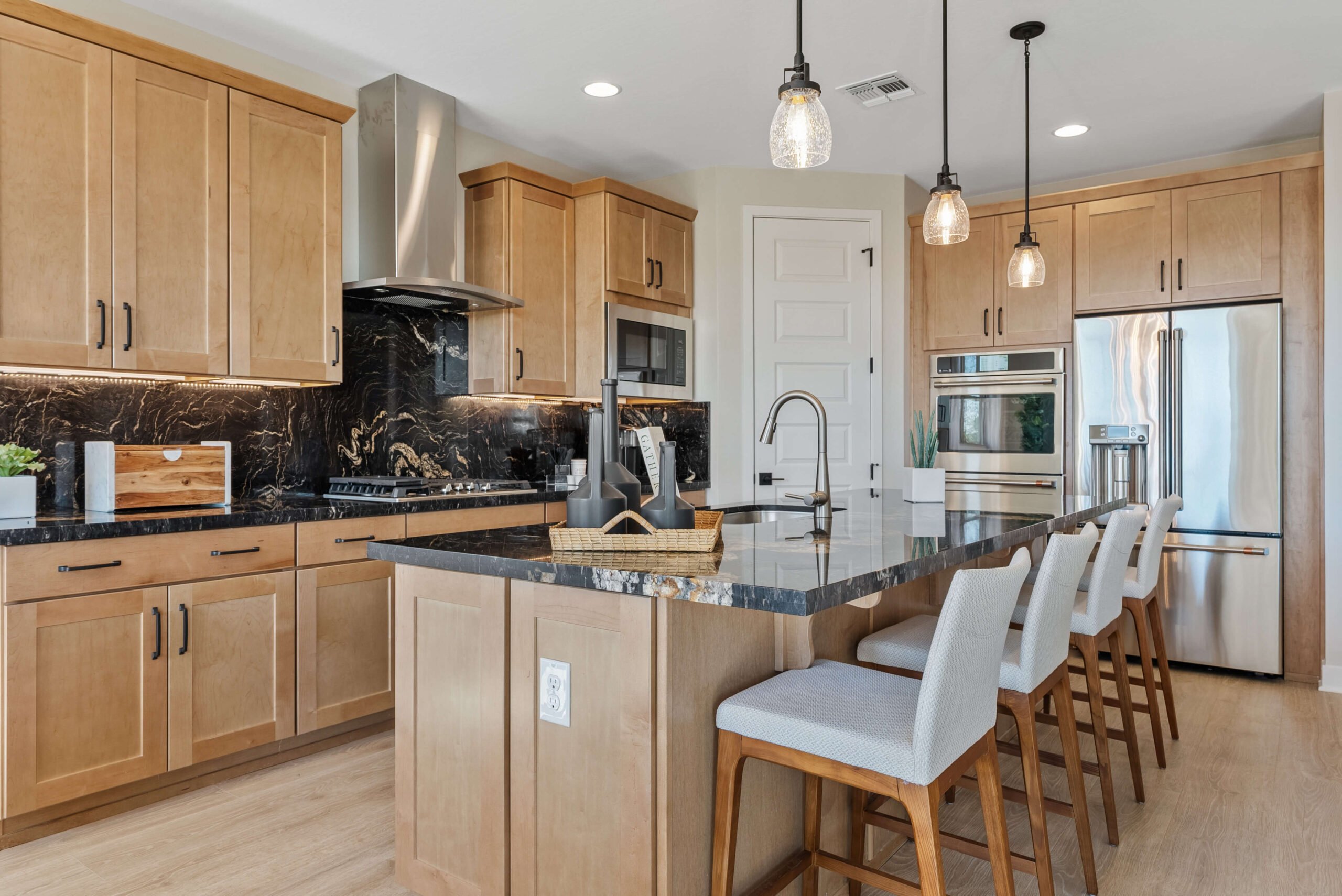 Modern kitchen with wooden cabinets, black marble countertops, island with four stools, pendant lights, stainless steel appliances, and a marble backsplash.