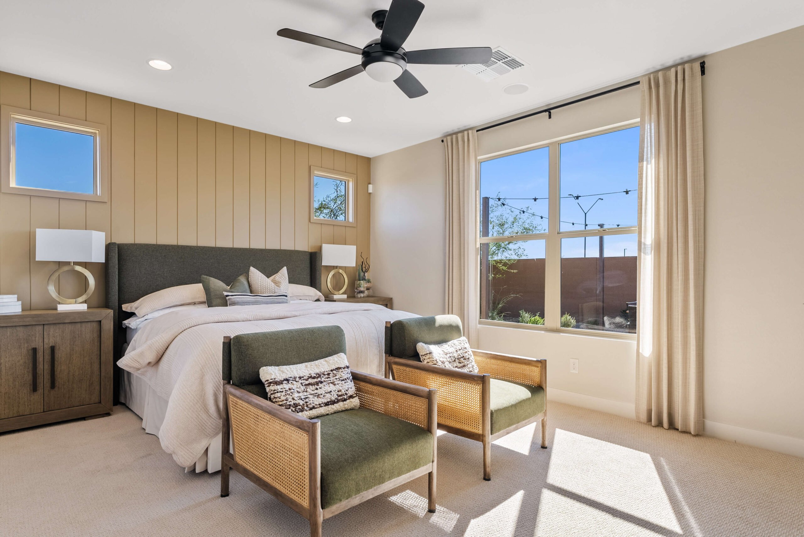 Bedroom with a large bed, two green armchairs, a ceiling fan, and a large window. Neutral tones and wood accents create a cozy, modern atmosphere.